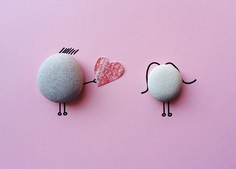 Two gray stones with drawn legs and a red heart between them on pink background