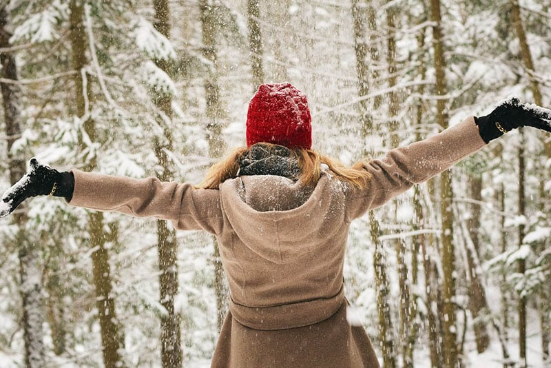 Woman in winter coat and red hat with arms outstretched in snowy woods