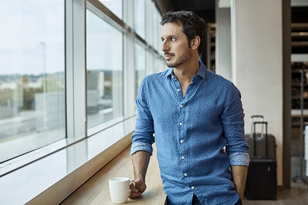 Thoughtful man in a blue shirt holding a coffee cup by a window