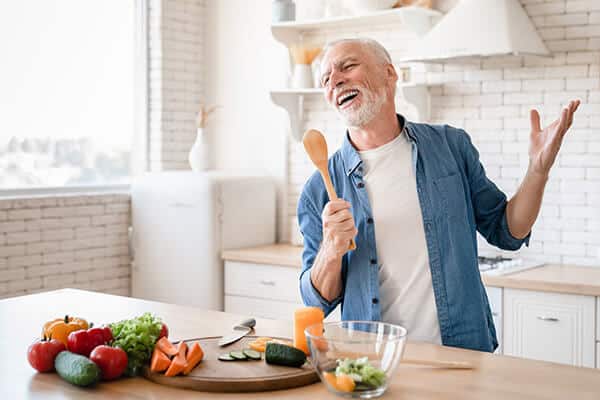 Laughing older man using a wooden spoon as a microphone in a kitchen