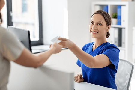 Receptionist in blue scrubs handing a card to a patient at a check-in desk